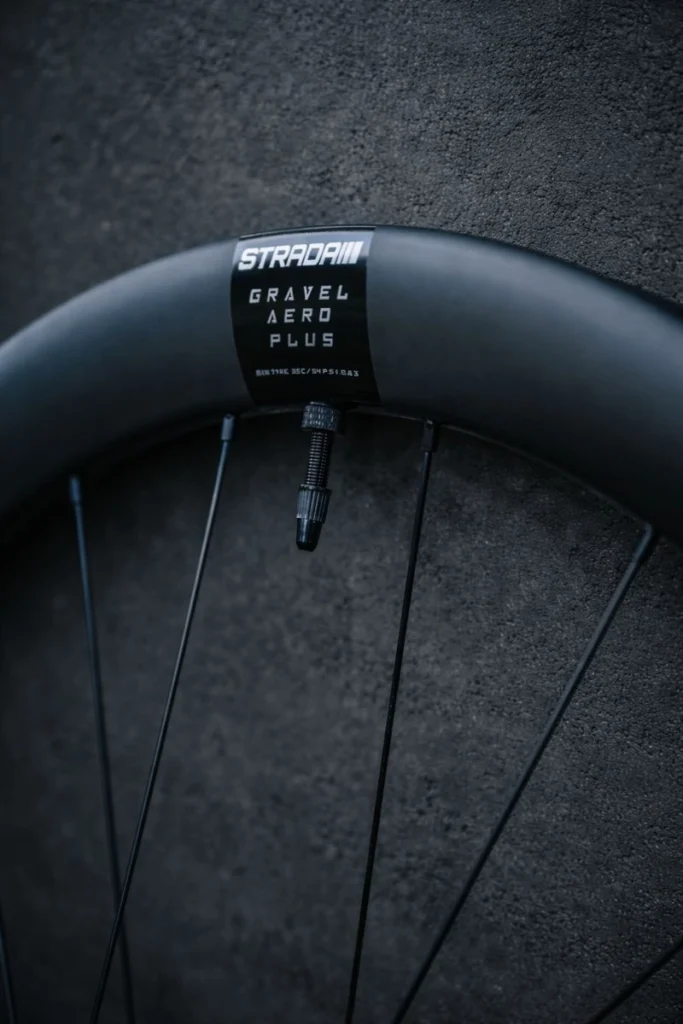 Close-up of a black carbon bicycle rim showing the Strada Gravel Aero Plus label, valve stem and radiating spokes against a dark textured background