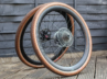 Two carbon gravel bike wheels with tan-wall tyres are propped against a dark wooden fence on a wooden deck. The front wheel partially overlaps the rear, showing the rear cassette and disc brake rotor.